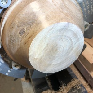 K&ouml;nnte beinhalten: Eine Holzsch&uuml;ssel, die auf einer Drehbank gedreht wird. Die Sch&uuml;ssel ist hellbraun mit einem dunkleren braunen Fleck in der Mitte. Die Drehbank ist aus Metall und hat einen schwarzen Sockel. Auf dem Sockel der Drehbank befinden sich Holzsp&auml;ne.
