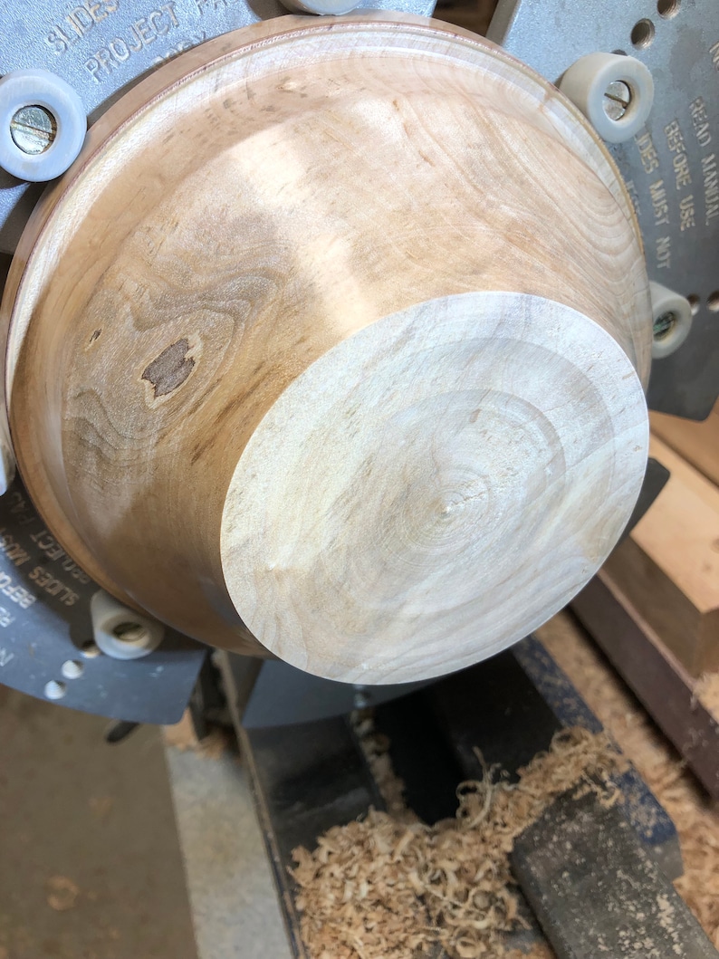 K&ouml;nnte beinhalten: Eine Holzsch&uuml;ssel, die auf einer Drehbank gedreht wird. Die Sch&uuml;ssel ist hellbraun mit einem dunkleren braunen Fleck in der Mitte. Die Drehbank ist aus Metall und hat einen schwarzen Sockel. Auf dem Sockel der Drehbank befinden sich Holzsp&auml;ne.