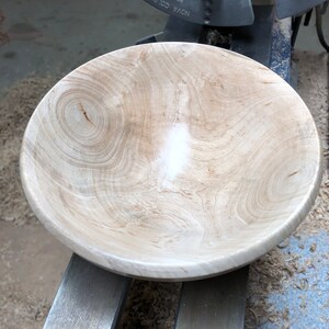 K&ouml;nnte beinhalten: Eine gro&szlig;e, unfertige Holzschale in hellbrauner Farbe mit einer glatten, polierten Oberfl&auml;che. Die Schale steht auf einer Drehbank, und um sie herum liegen Holzsp&auml;ne auf dem Boden.