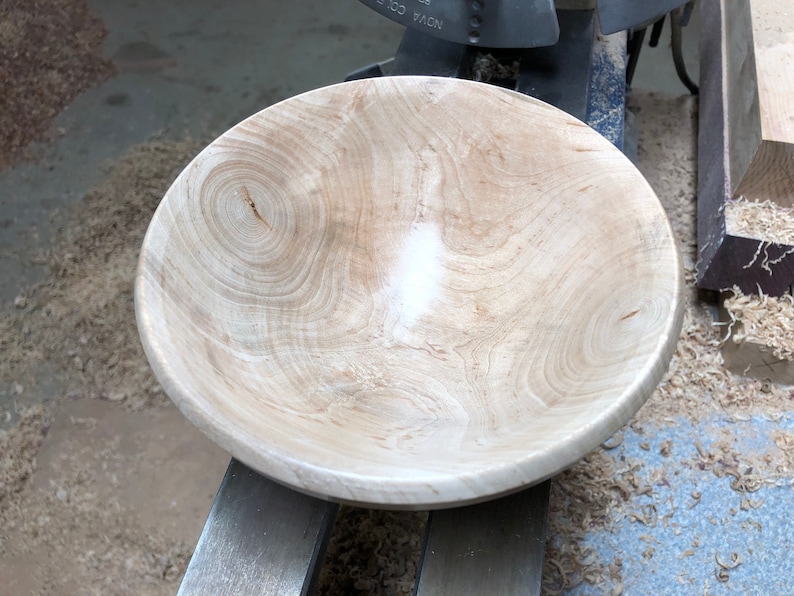 K&ouml;nnte beinhalten: Eine gro&szlig;e, unfertige Holzschale in hellbrauner Farbe mit einer glatten, polierten Oberfl&auml;che. Die Schale steht auf einer Drehbank, und um sie herum liegen Holzsp&auml;ne auf dem Boden.