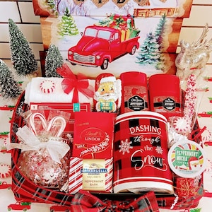 May include: A Christmas-themed gift basket with a red and black plaid pattern. It contains a mug with "Dashing through the Snow" text, chocolates, candy canes, and other festive items. A sign that says "Home for the Holidays" is in the background.