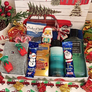 May include: A Christmas gift basket filled with various treats and toiletries. Includes a gray sweater, chocolate bars, candy, body spray, and a bottle of hair and body wash. The basket is decorated with festive wrapping and ribbons.