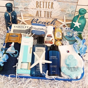 May include: A gift basket with a beach theme, featuring blue and white items. Includes a blue towel with a sailboat, bath products, chocolates, and a bath bomb. The background has a sign that says "Better at the Beach".