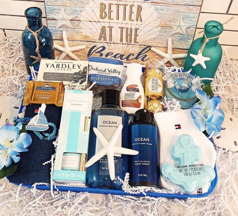 May include: A gift basket with a beach theme, featuring blue and white items. Includes a blue towel with a sailboat, bath products, chocolates, and a bath bomb. The background has a sign that says "Better at the Beach".