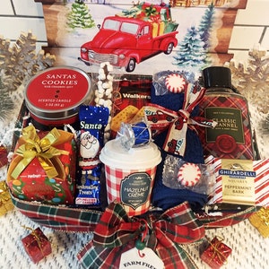 May include: A Christmas-themed gift basket filled with festive treats and toiletries. Includes a red candle, chocolates, cookies, a coffee cup, and a bottle of body wash. The basket is decorated with a plaid ribbon and small gift boxes.