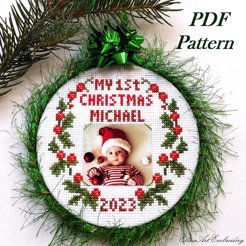 Puede incluir: Un adorno de punto de cruz blanco con una corona de acebo verde, bayas rojas y un marco de fotos. El texto "My 1st Christmas Michael 2023" est&aacute; bordado en hilo rojo.