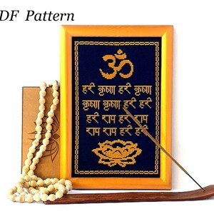 May include: A framed cross-stitch pattern featuring the Sanskrit word Om, a lotus flower, and a mantra in Hindi. The frame is made of wood and has a gold finish. The pattern is on a dark blue fabric with gold thread.