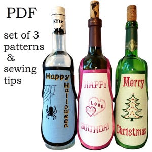 May include: Set of three wine bottle covers with cross-stitch designs. The covers are blue, pink, and green with the words "Happy Halloween", "Happy Birthday", and "Merry Christmas" respectively.