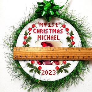 Puede incluir: Un adorno navide&ntilde;o redondo y blanco bordado con hojas de acebo verdes y bayas rojas. El adorno tiene un marco de fotos con un beb&eacute; con un atuendo a rayas rojas y blancas. El texto "MY 1st CHRISTMAS MICHAEL 2023" est&aacute; bordado en el adorno. El adorno est&aacute; decorado con oropel verde.