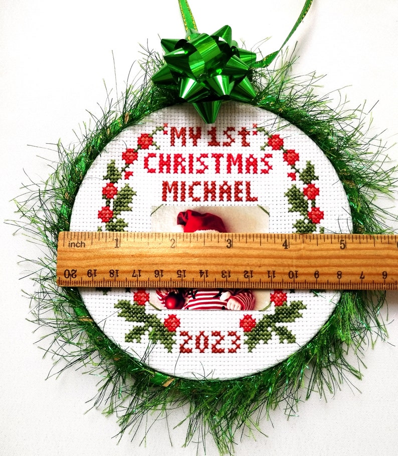 Puede incluir: Un adorno navide&ntilde;o redondo y blanco bordado con hojas de acebo verdes y bayas rojas. El adorno tiene un marco de fotos con un beb&eacute; con un atuendo a rayas rojas y blancas. El texto "MY 1st CHRISTMAS MICHAEL 2023" est&aacute; bordado en el adorno. El adorno est&aacute; decorado con oropel verde.