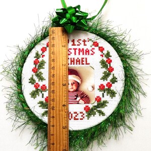 Puede incluir: Un adorno navide&ntilde;o redondo y blanco bordado con un borde de acebo verde y un dise&ntilde;o de bayas rojas. El adorno tiene una foto de un beb&eacute; con un gorro de Pap&aacute; Noel y el texto "1&ordf; Navidad Michael 2023". El adorno est&aacute; decorado con oropel verde.
