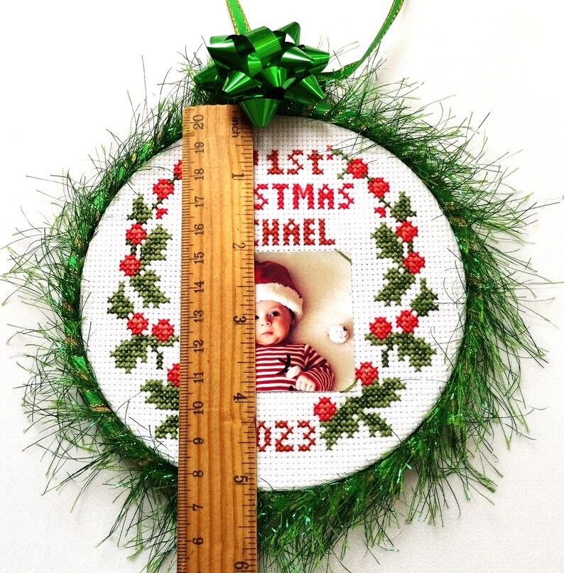 Puede incluir: Un adorno navide&ntilde;o redondo y blanco bordado con un borde de acebo verde y un dise&ntilde;o de bayas rojas. El adorno tiene una foto de un beb&eacute; con un gorro de Pap&aacute; Noel y el texto "1&ordf; Navidad Michael 2023". El adorno est&aacute; decorado con oropel verde.