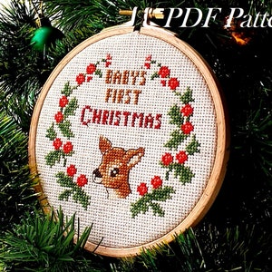 以下が含まれることがあります： 赤ちゃん最初のクリスマスのクロスステッチデザインが施された白い刺繍枠。鹿とヒイラギのベリーが描かれています。テキストは「Baby's First Christmas」です。