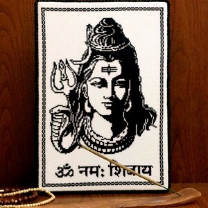 May include: Black and white cross-stitch embroidery of the Hindu deity Shiva with the Sanskrit phrase "Om Namah Shivaya" below. The embroidery is framed and displayed on a wooden surface with an incense stick.