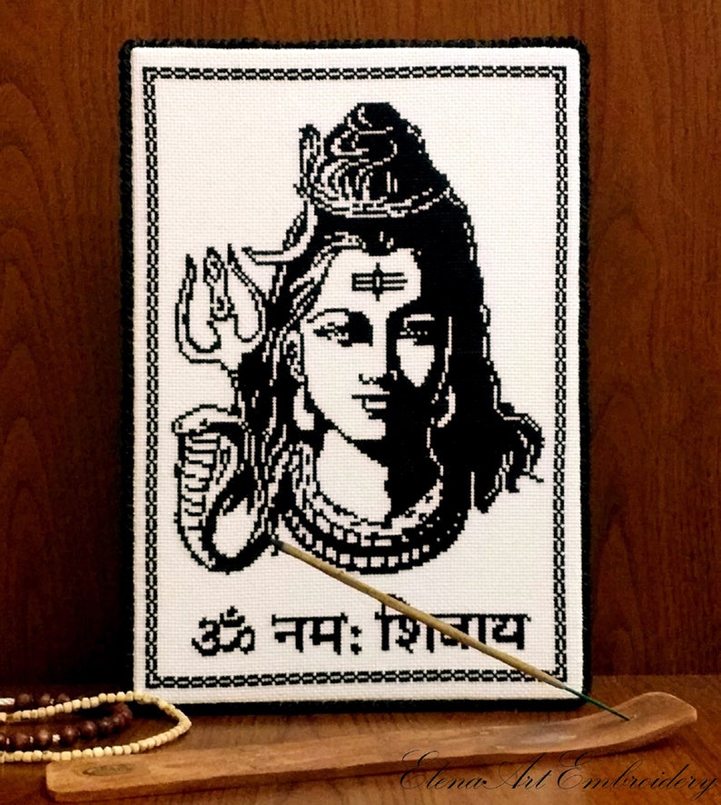 May include: Black and white cross-stitch embroidery of the Hindu deity Shiva with the Sanskrit phrase "Om Namah Shivaya" below. The embroidery is framed and displayed on a wooden surface with an incense stick.