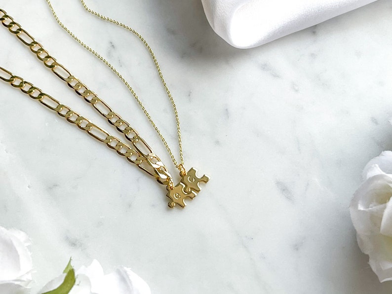 May include: Two gold necklaces with a puzzle piece pendant. The necklaces are layered on a white marble surface.