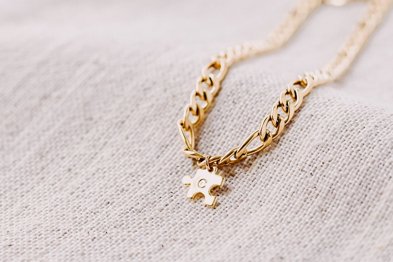 May include: A gold chain necklace with a gold puzzle piece charm. The puzzle piece has the letter "C" engraved on it.