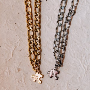 May include: Two gold and silver chain necklaces with puzzle piece pendants. The gold chain has a "C" on the pendant and the silver chain has a "B" on the pendant.