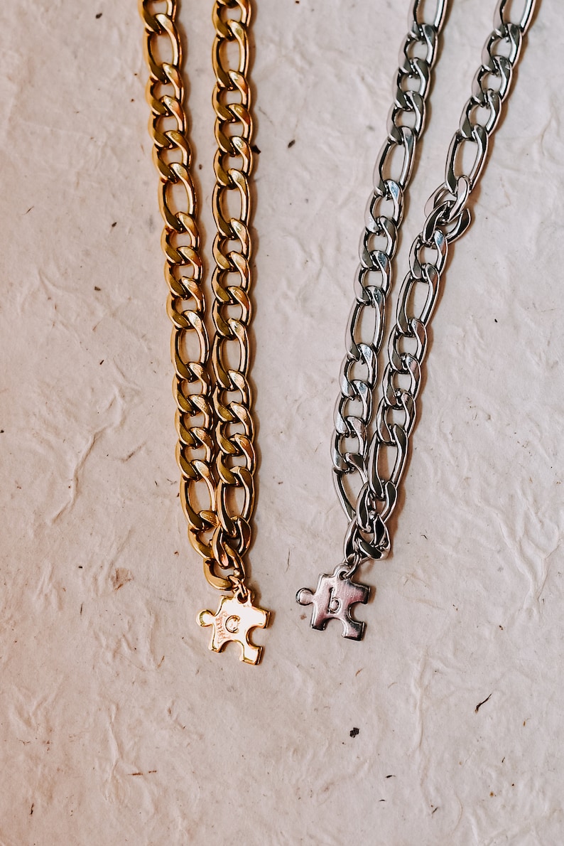 May include: Two gold and silver chain necklaces with puzzle piece pendants. The gold chain has a "C" on the pendant and the silver chain has a "B" on the pendant.