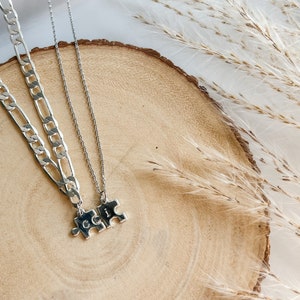 May include: Two silver necklaces with puzzle piece pendants. The necklaces are on a wooden surface with dried grass in the background.