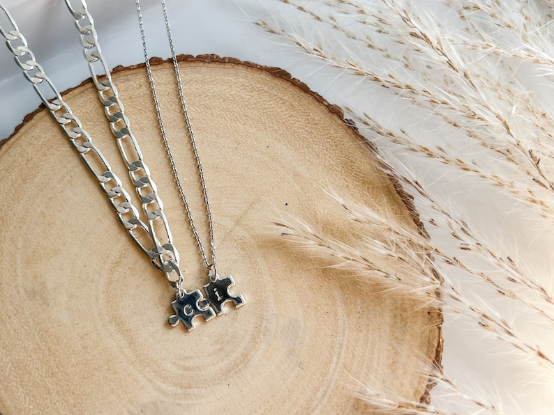 May include: Two silver necklaces with puzzle piece pendants. The necklaces are on a wooden surface with dried grass in the background.