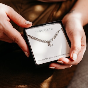May include: A silver chain necklace with a puzzle piece charm in a white box with the text "JUNE SIXTH" and "You are the piece that completes me".