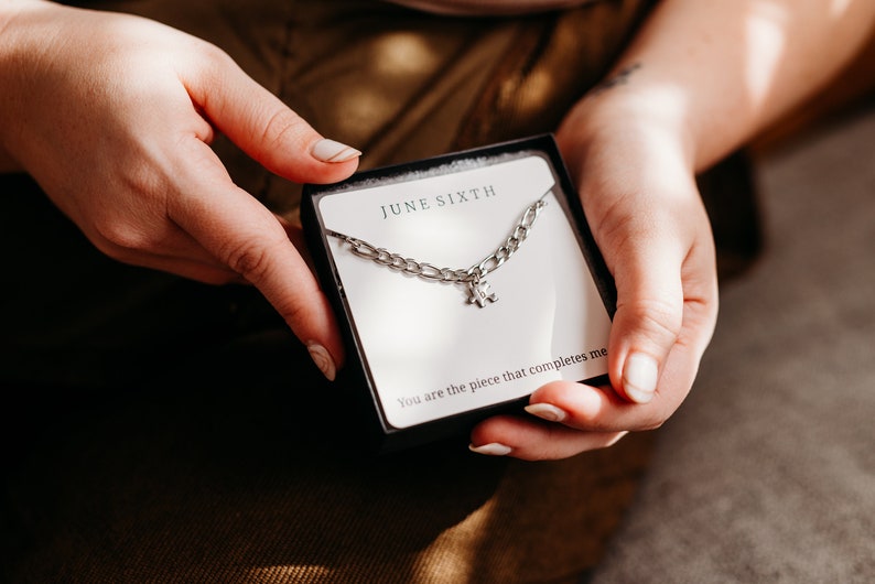 May include: A silver chain necklace with a puzzle piece charm in a white box with the text "JUNE SIXTH" and "You are the piece that completes me".