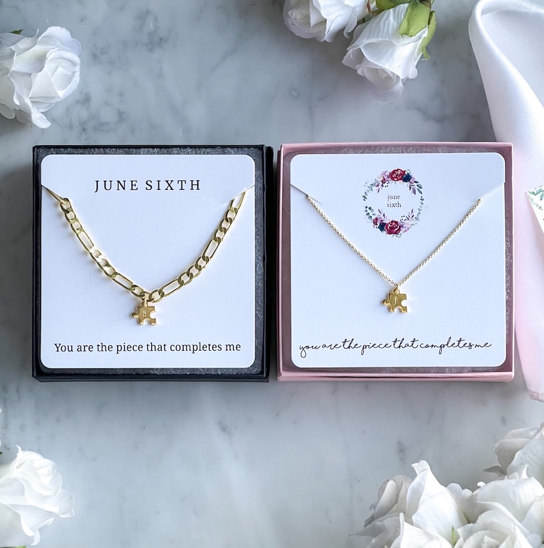May include: Two gold necklaces in black and pink boxes. The necklaces feature a small gold puzzle piece charm. The boxes have the text "JUNE SIXTH" and "You are the piece that completes me".