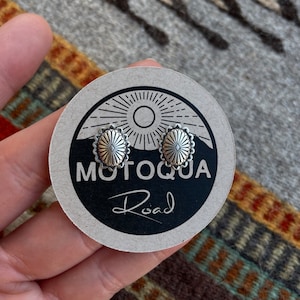May include: A pair of silver-colored stud earrings with a floral design, presented on a round card with the text "MOTOQUA Road". The earrings are small and detailed, with a textured surface. The card is held against a patterned textile background.
