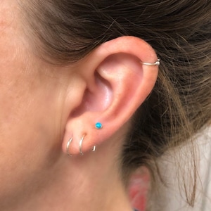 May include: Close-up of an ear with multiple earrings. The ear has a small turquoise stud, two silver hoops, and a silver hoop earring on the upper ear. The skin is fair-toned.