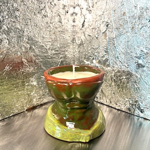 May include: A ceramic candle vessel shaped like a muscular torso with defined abdominal muscles. The vessel is glazed in shades of olive green and brown with a reddish-brown rim. A white candle wick is centered in the off-white wax filling the top.