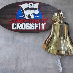 May include: A brass ship's bell with a braided rope and a wooden sign. The sign has the text "COME AND TAKE IT CROSSFIT" with a flag and cannon design. The sign also has the text "45LB" and "20KG".