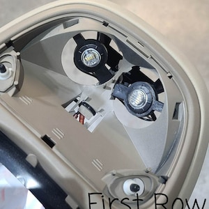 May include: Close-up of a beige car interior dome light with two white LED bulbs. The light is in a recessed housing with a black plastic frame. The text "First Row" is visible in the lower right corner of the image.