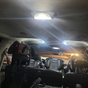 May include: The interior of a vehicle featuring multiple black Graco child safety seats installed in the back rows. Bright overhead dome lights illuminate the cabin, casting a glow over the dark upholstery and the folded stroller in the foreground.