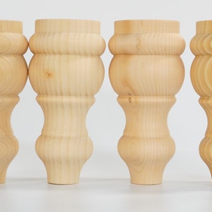May include: Four unfinished wooden furniture legs with a turned design. The legs are made of light wood and have a smooth finish.
