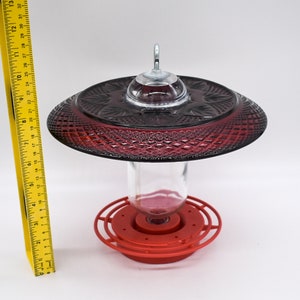 May include: Red glass hummingbird feeder with a clear glass bottle and a red plastic base. The feeder has a decorative, diamond-shaped pattern.