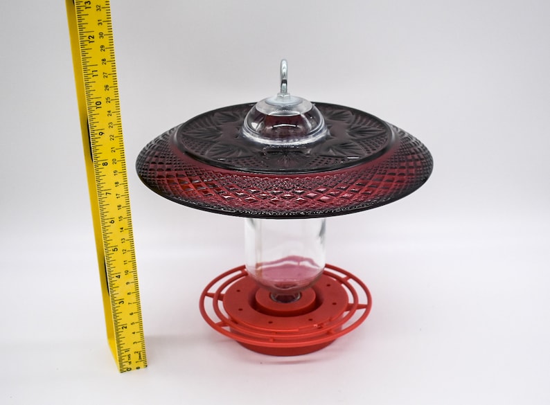 May include: Red glass hummingbird feeder with a clear glass bottle and a red plastic base. The feeder has a decorative, diamond-shaped pattern.