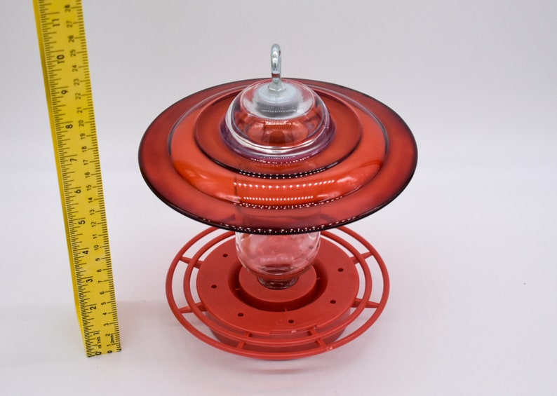 A red hummingbird feeder with a clear glass reservoir and a red plastic base. The feeder has a clear plastic dome with a metal hook on top. The feeder is designed to attract hummingbirds.