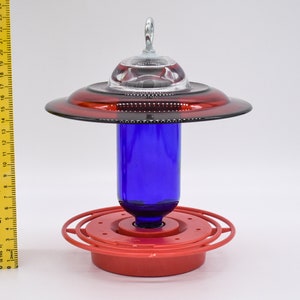 May include: A red and cobalt blue glass hummingbird feeder with a clear glass dome on top. The feeder has a Best 1 red base and a red ring around the middle.