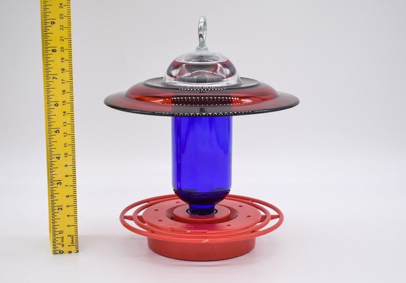 May include: A red and cobalt blue glass hummingbird feeder with a clear glass dome on top. The feeder has a Best 1 red base and a red ring around the middle.