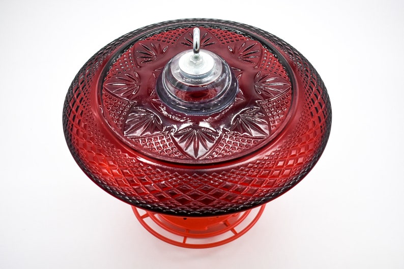 May include: A red glass hummingbird feeder with a decorative, patterned top and a clear plastic feeding area. The feeder has a metal hook for hanging.