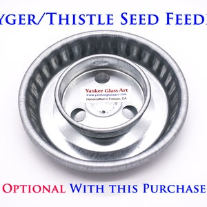 May include: A silver metal bird feeder for nyger or thistle seeds. The feeder has a round base with a smaller, round, inner ring. The inner ring has three holes for birds to perch on. The feeder is labeled "Yankee Glass Art" and "Handcrafted in Folsom, CA".