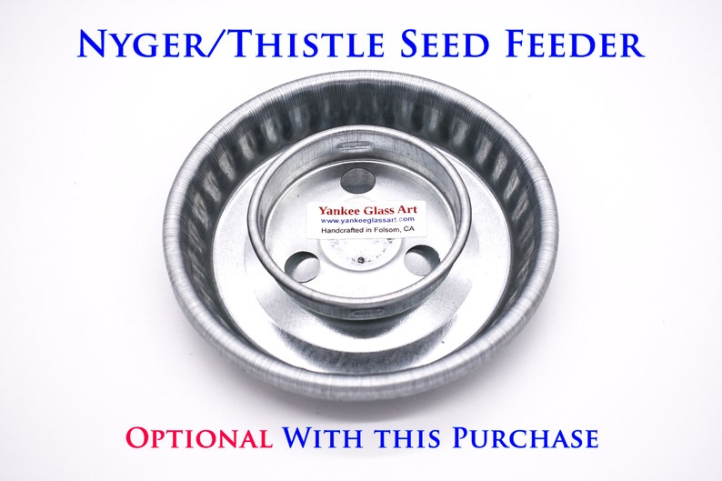 May include: A silver metal bird feeder for nyger or thistle seeds. The feeder has a round base with a smaller, round, inner ring. The inner ring has three holes for birds to perch on. The feeder is labeled "Yankee Glass Art" and "Handcrafted in Folsom, CA".