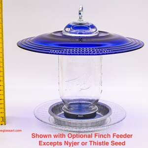May include: A blue glass bird feeder with a clear glass jar and a metal base. The feeder is shown with an optional finch feeder. "Shown with Optional Finch Feeder Excepts Nyjer or Thistle Seed" is written below the feeder.