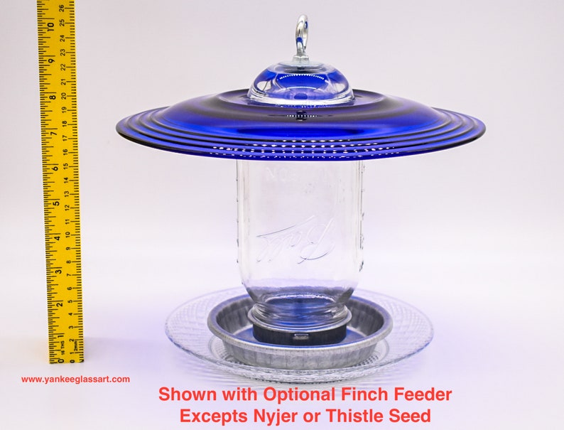 May include: A blue glass bird feeder with a clear glass jar and a metal base. The feeder is shown with an optional finch feeder. "Shown with Optional Finch Feeder Excepts Nyjer or Thistle Seed" is written below the feeder.