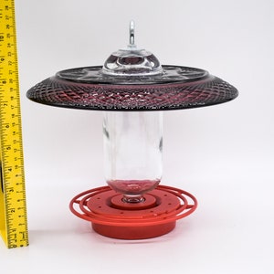 May include: A red hummingbird feeder with a clear glass bottle and a dark red, glass, decorative top. The feeder has a metal ring around the bottle and a red base.