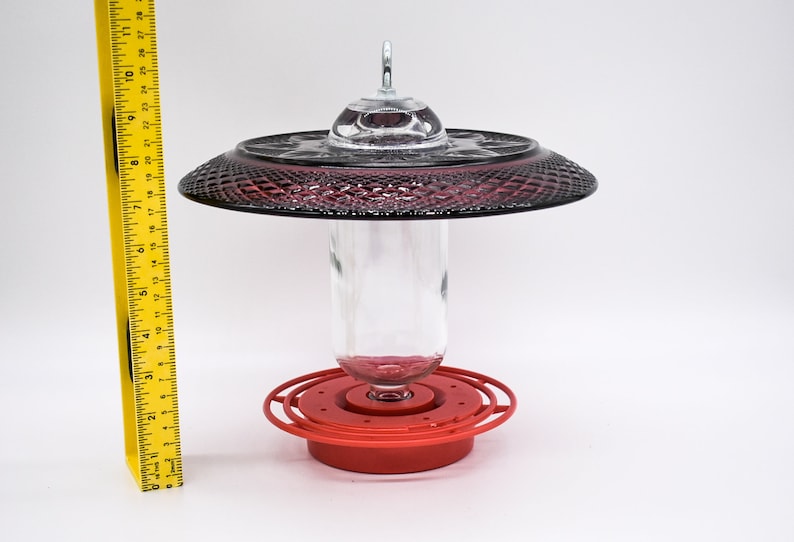 May include: A red hummingbird feeder with a clear glass bottle and a dark red, glass, decorative top. The feeder has a metal ring around the bottle and a red base.