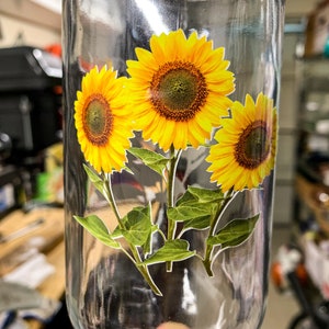 May include: A clear glass bottle with a sticker of three yellow sunflowers with green leaves.