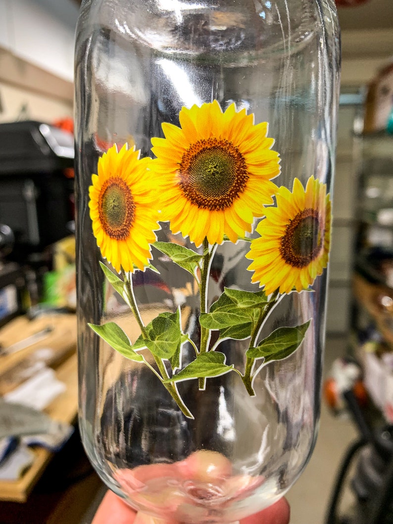 May include: A clear glass bottle with a sticker of three yellow sunflowers with green leaves.