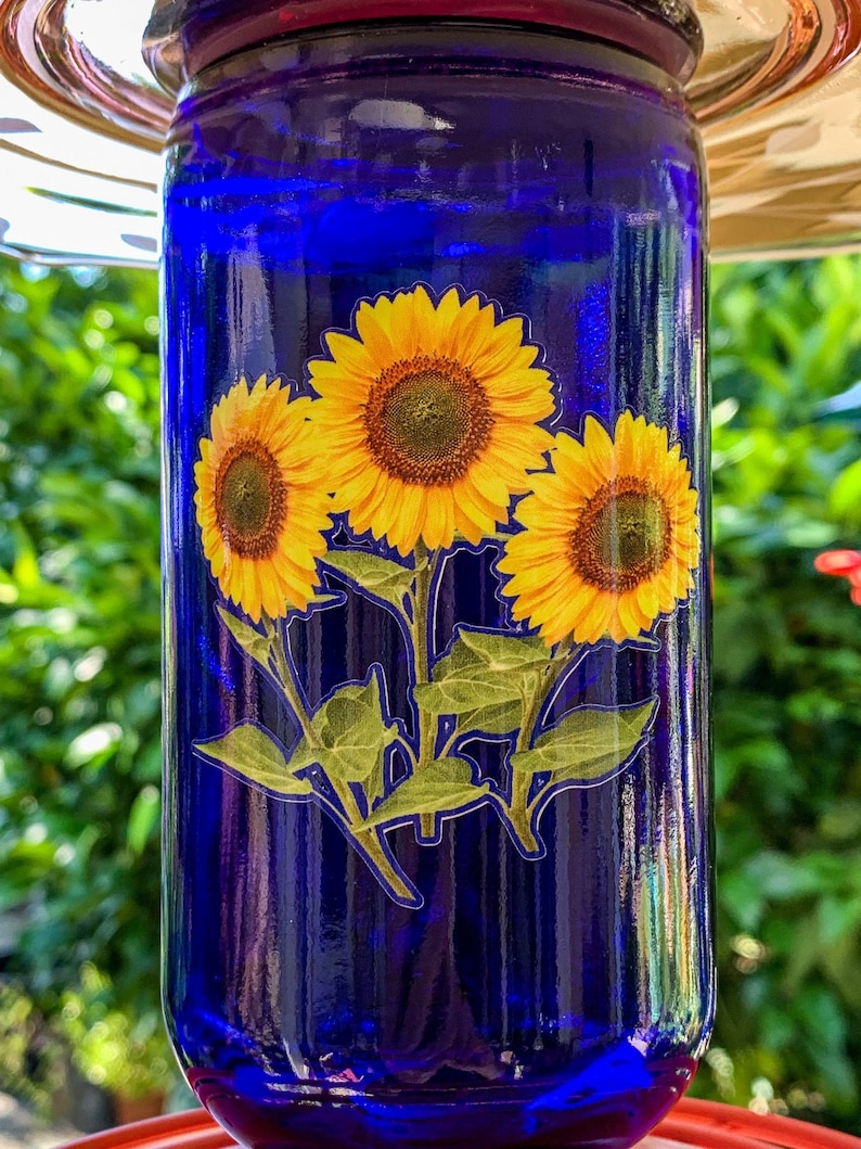 May include: A blue glass hummingbird feeder with a sticker of three yellow sunflowers with green leaves and stems.